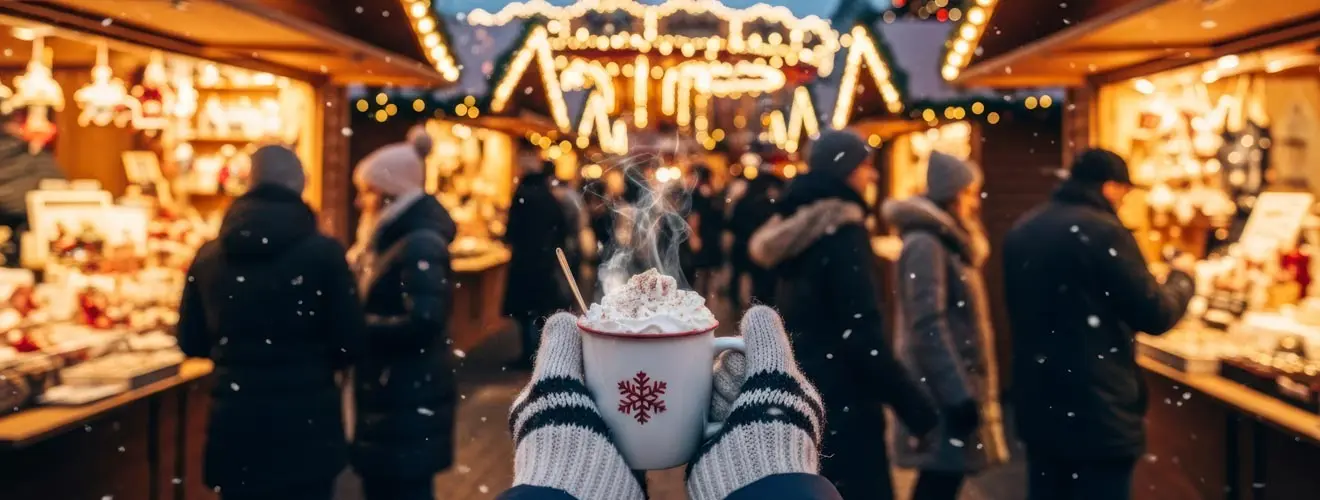 Hände in Winterhandschuhen halten einen dampfenden Becher vor beleuchteten Weihnachtsmarktständen